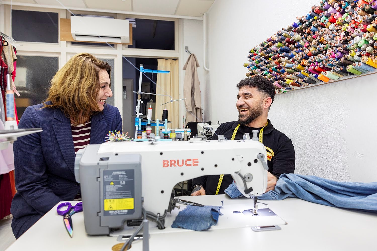Nermina Kundić zit naast een medewerker van De Diamanten Schaar aan tafel met een naaimachine op de voorgrond. 