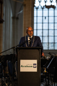 Burgemeester Marcouch geeft zijn toespraak tijdens de Dodenherdenking. Hij staat achter een lessenaar met het logo van de gemeente Arnhem.